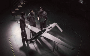 Three individuals in camouflage uniforms discuss around a model drone in a dimly lit room with a staircase and monitor. Yellow-black striped lines mark the floor. The image relates to a "Mobile App Development Cost Guide for Defense Contractors," focusing on military technology and development.