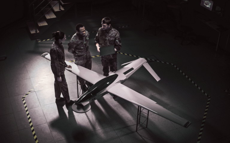 Three individuals in camouflage uniforms discuss around a model drone in a dimly lit room with a staircase and monitor. Yellow-black striped lines mark the floor. The image relates to a "Mobile App Development Cost Guide for Defense Contractors," focusing on military technology and development.