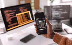 A hand holding a smartphone displaying code, with computer screens in the background showing programming interfaces.