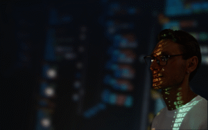 A man in a white shirt with code projected onto his face and neck, standing against a dark background with blurred digital displays, symbolizing mobile application development systems for defense and space. The image highlights advanced tech integration for secure, innovative solutions in these sectors.