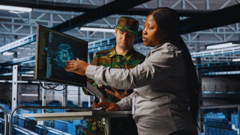 Two people, one in military uniform, collaborating at a workstation with a screen displaying AI data in an industrial setting.