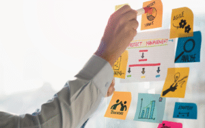 Image of a hand writing "Project Management" on a whiteboard with sticky notes labeled Agile, Analyze, Strategy, Teamwork, and Market Share, illustrating project management concepts.